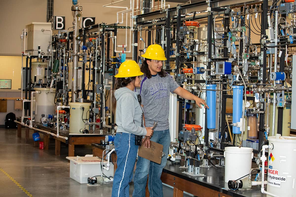 Two San Juan students in yellow hard hats look at a clipboard with machinery in the background