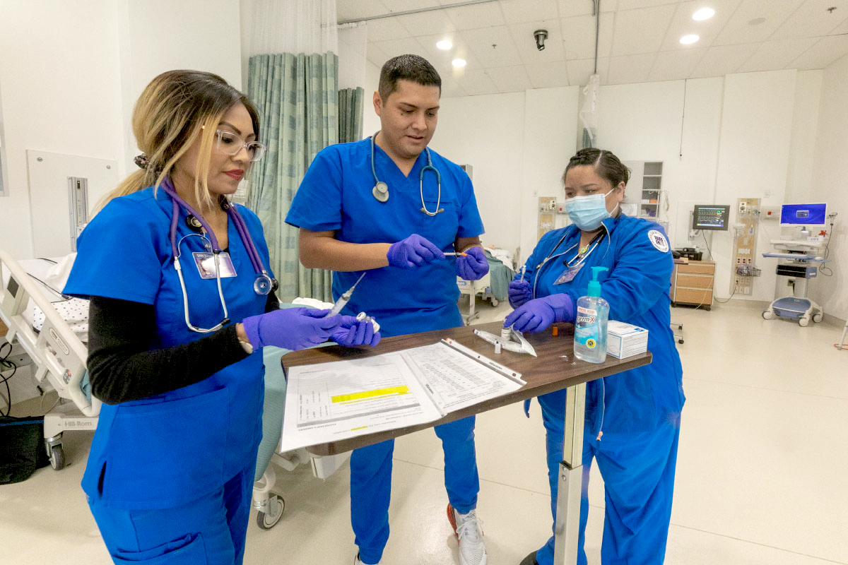 Three students wearing scrubs, working in a hospital room.
