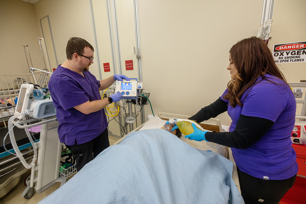 Two students stand next to a hospital bed, working on simulated patient.